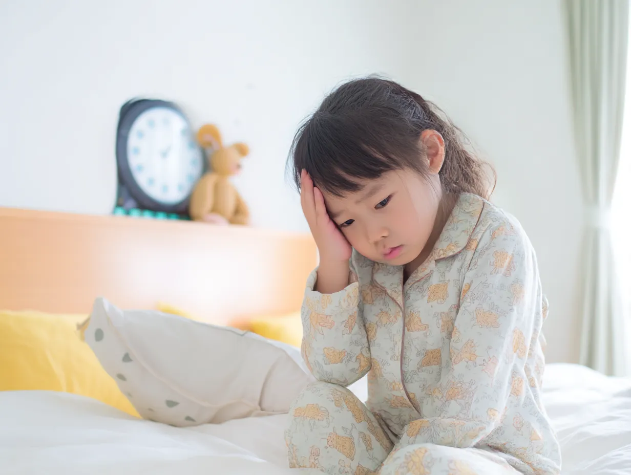 幼い女の子の発熱写真｜寝起きのパジャマ姿の女の子が頭を押さえ、不調を訴えるような表情を見せるシーン。柔らかい布団と優しい色合いが家庭的で安心感のある雰囲気を与える。