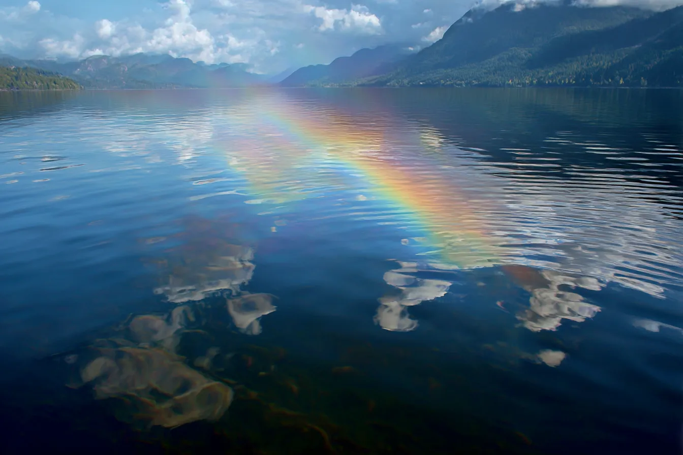 レインボーの反射が映る湖の背景｜湖面に映る虹とその反射が、美しい自然の背景を形成しています。空と湖が溶け合うように、虹がその上に広がり、穏やかな水面に柔らかな色を映し出しています。