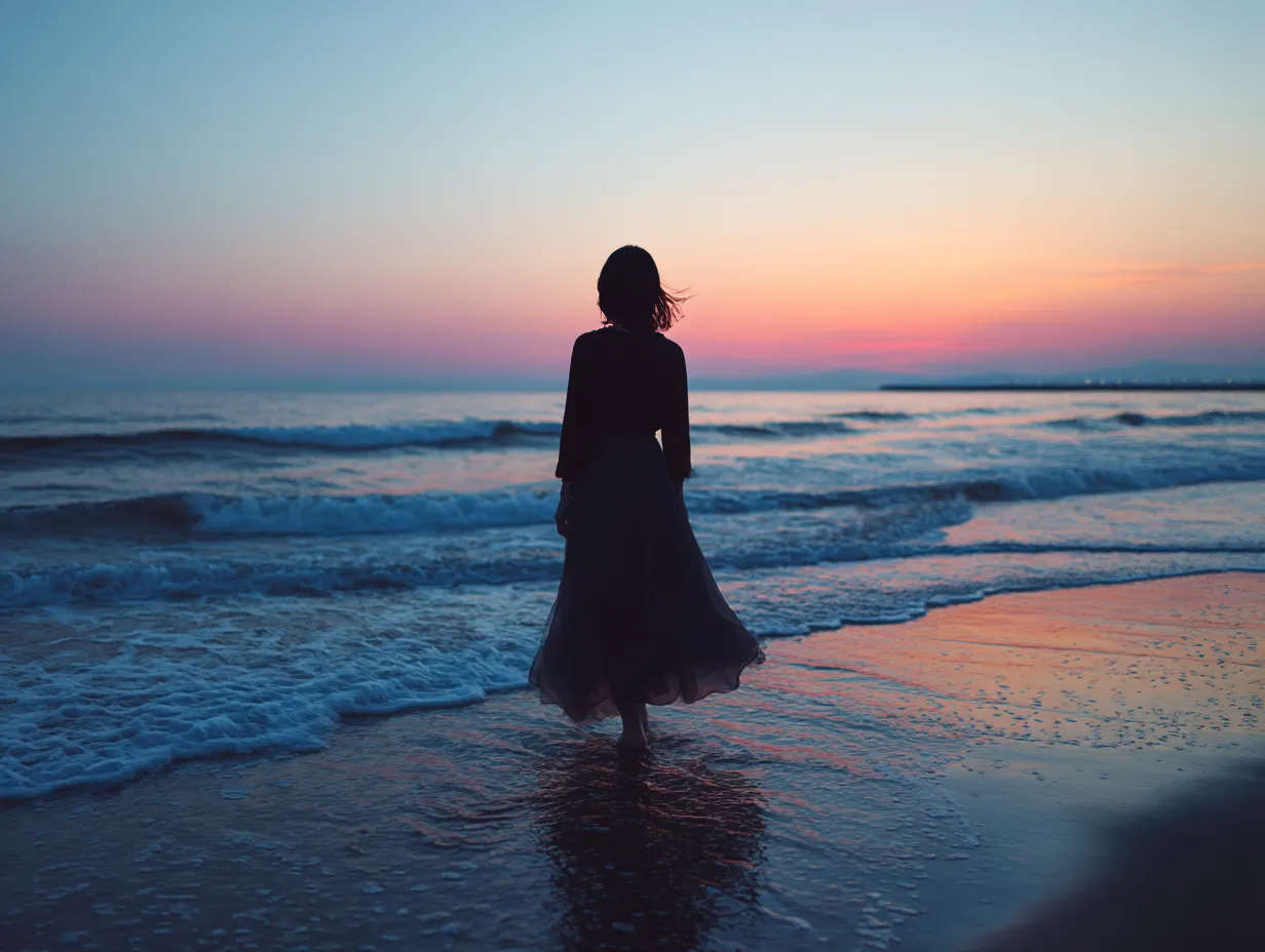 夕暮れの浜辺に佇む後ろ姿の女性の画像｜柔らかな夕焼けのグラデーションが海を染める中、波打ち際に立つ後ろ姿の女性の画像。ロングスカートの揺らぎとシルエットが幻想的な印象を与え、静寂と余韻を感じさせる構図。