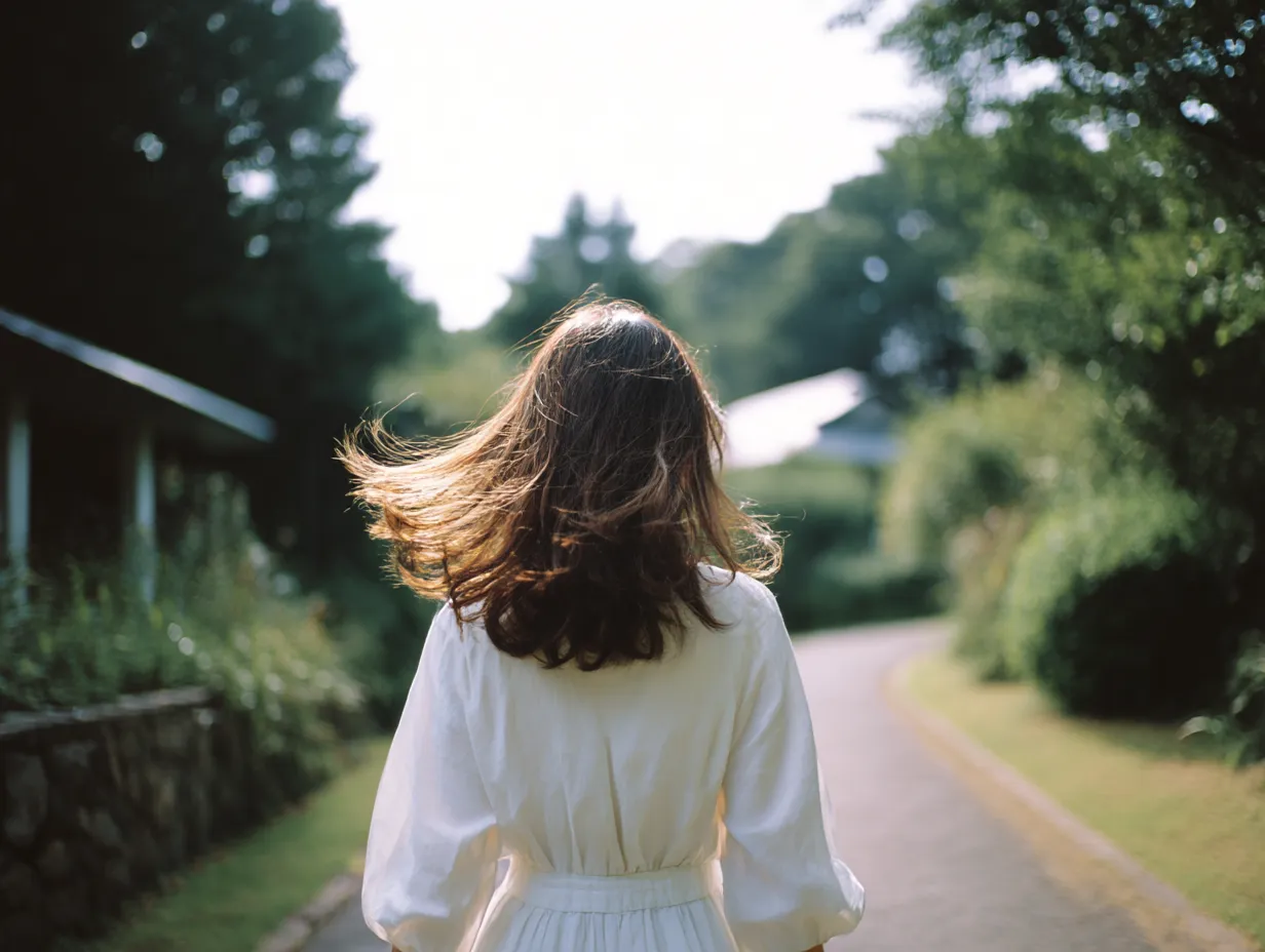 緑道を歩く白いワンピースの後ろ姿の女性の画像｜明るい自然光の中、緑に囲まれた道を歩く白いワンピースの後ろ姿の女性の画像。柔らかい逆光で髪が透け、初夏の穏やかな空気感と爽やかな透明感を感じる描写。