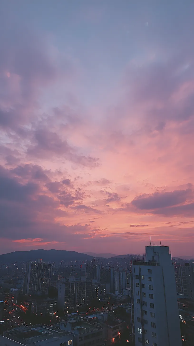 紫エモい韓国壁紙 夕映え空｜都市のビル群を背景に、雲間から広がる紫とピンクの夕焼けが空を染める。紫エモい韓国壁紙として、光と影の対比が感情的な余韻を引き立てる。
